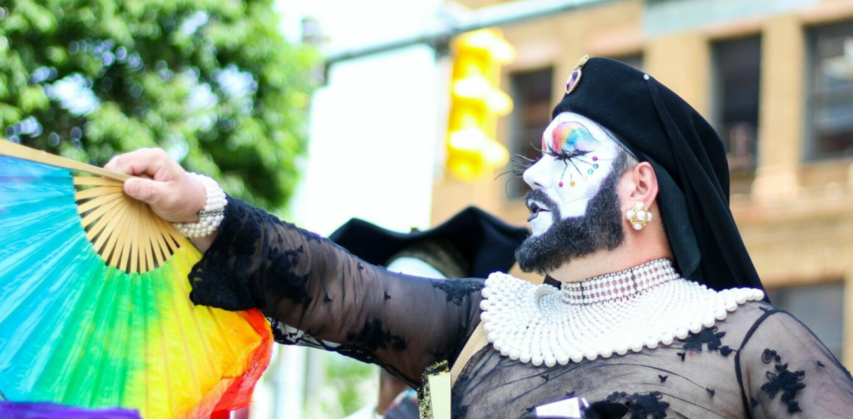 Persona caracterizada con barba portando un abanico con los colores de la bandera gay